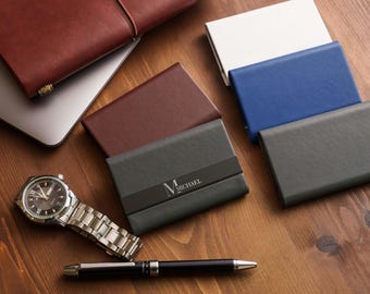 May include: A collection of rectangular card holders in various colors including dark gray, brown, blue, and white. One dark gray card holder features the name MICHAEL printed in white. A silver wristwatch and a black pen are also present.