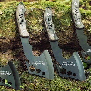 May include: A collection of personalized axes with wooden handles and dark gray blades. Each axe features a name engraved on the handle and the text "Groomfiman 10.21.2023" on the blade. The axes are displayed on a mossy surface.