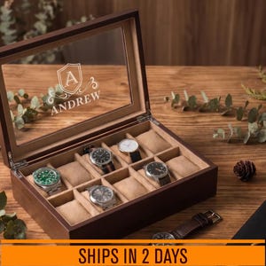 May include: A dark brown wooden watch display box with a glass lid featuring a white engraved shield emblem and the name Andrew. The interior is lined with beige fabric and contains multiple compartments with cushions holding various wristwatches.