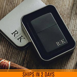 May include: A matte black metal lighter with the engraved initials R|K, presented in a matching silver-toned metal gift tin that also features the engraved initials R|K on the lid. The lighter rests inside a black velvet-lined insert within the tin.