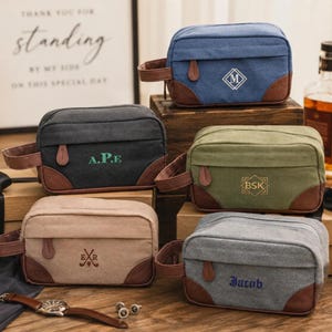 Puede incluir: Una colección de cinco neceseres rectangulares de lona con detalles en piel sintética marrón y asas laterales. Cada bolsa presenta monogramas o nombres bordados únicos, incluyendo M, A.P.E, BSK, E R y Jacob, en varios colores de hilo.