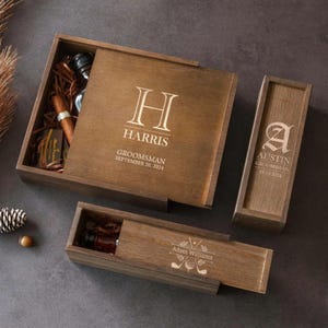 May include: Three dark brown wooden boxes with sliding lids. The large square box is engraved with H, HARRIS, GROOMSMAN, SEPTEMBER 20, 2024. The tall rectangular box features A, AUSTIN, GROOMSMAN, 09.20.2024. The long box shows Adam Williams and golf icons.