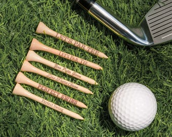 Può includere: Sei tee da golf in legno di colore chiaro con testo stampato, una pallina da golf bianca con fossette e una testa di mazza da golf in metallo. I tee presentano le frasi LET'S PAR TEE, Zero Putts Given e PERFECT SWING in vari caratteri.