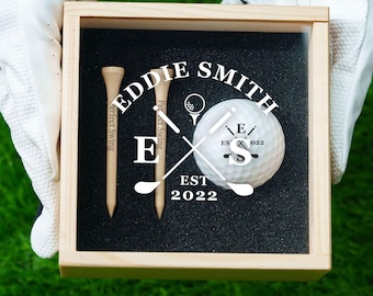 May include: A light-colored wooden box containing two wooden golf tees and one white golf ball, all set in black foam. The glass lid features white printed text reading EDDIE SMITH, E S, and EST 2022, along with a graphic of crossed golf clubs.