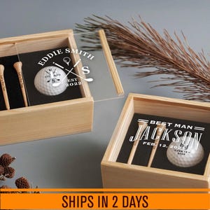 Peut inclure: Deux boîtes en bois clair avec couvercles coulissants transparents, contenant chacune une balle de golf blanche et deux tees en bois. La boîte de gauche porte le texte Eddie Smith Est 2022, celle de droite Best Man Jackson Feb 12 2025.