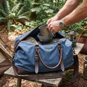 May include: A blue canvas duffel bag with brown leather trim and handles, featuring the name Abigail embroidered in dark thread on the front. The bag is open, revealing metallic camping mugs inside, and includes a detachable shoulder strap.