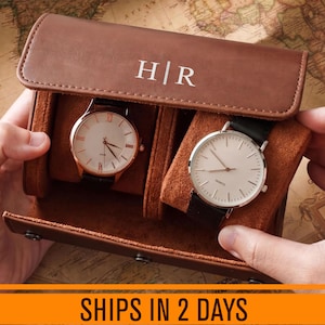 May include: A brown leather watch roll case featuring the monogram H|R on the flap, containing two wristwatches with round faces, black leather straps, and white dials. The interior is lined with soft brown suede, and the case secures with metal snaps.