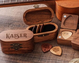 May include: A wooden guitar pick box with the name KAISER engraved on the lid, featuring a metal latch. Beside it, an open wooden box contains three picks, and another small box holds a pick engraved with P.N. Two loose picks are nearby.