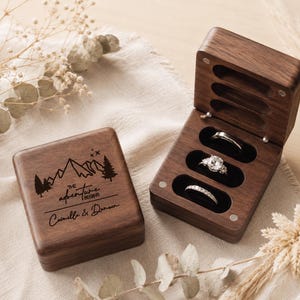 May include: A dark brown wooden ring box with a hinged lid featuring a laser-engraved mountain landscape, pine trees, and the text "THE ADVENTURE BEGINS" with the names "Camella & Danson". The open box contains three slots holding three rings.