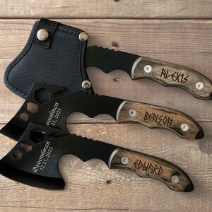 May include: Three black metal hatchets with wooden handles, each engraved with a name in a runic-style font. Two hatchets feature the text 'Groomsman 12 21, 2023' on the blade, while one hatchet is stored in a black nylon sheath with silver snaps.