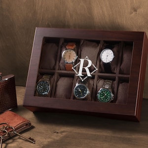 May include: A dark brown wooden watch display case with a glass lid featuring a white letter R logo, containing five wristwatches on brown cushions. Beside it sits a brown textured metal flask and a brown leather wallet with two metal keys.