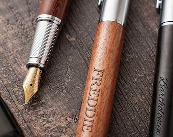 May include: Three pens with metallic accents. One fountain pen features a wood-grain barrel and a gold-toned nib marked Iridium Point. A second pen has a wood barrel engraved with the name FREDDIE. A third pen is black with the name Bartholomew.