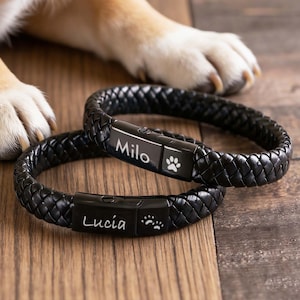 May include: Two black braided bracelets with rectangular metal clasps. One clasp is engraved with the name Milo and a single paw print, while the other is engraved with the name Lucia and two small paw prints. The bracelets have a woven texture.