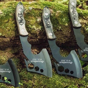 May include: Three black metal hatchets with wooden handles. Each handle is engraved with a name: CALEB, BRUKE, and MICHAEL. The blades feature hexagonal cutouts and the text "Groomsman 10 21, 2023" printed in white on the flat metal surface.