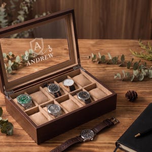 May include: A rectangular dark wood watch box with a glass lid featuring a white engraved shield emblem with the letter A and the name ANDREW. The interior is lined with beige fabric and contains ten individual compartments with watch pillows.