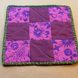 May include: A small purple and pink patchwork quilt with a green border. The quilt is made of squares of different fabrics with floral patterns. The quilt is quilted with a diamond pattern.