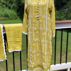 May include: A yellow embroidered dress with white lace trim and a matching yellow shalwar. The dress has a round neckline and a button-up front.