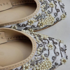 May include: A pair of white embroidered flat shoes with pearl embellishments. The shoes have a pointed toe and a low heel.