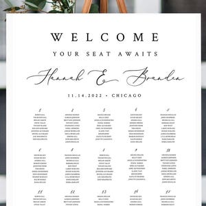 May include: A white wedding seating chart on an easel. The sign reads "WELCOME YOUR SEAT AWAITS" above the names "Hannah & Brandon" and the date and location. The sign is decorated with greenery and flowers.