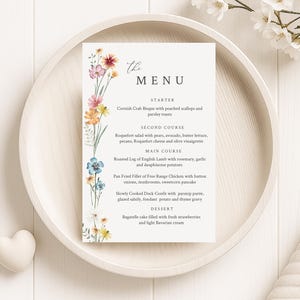 Può includere: Un biglietto del menu di nozze con accenti floreali ad acquerello. Il menu presenta un elenco di portate, tra cui antipasti, portate principali e dessert. Il biglietto è appoggiato su un vassoio di legno con una decorazione a forma di cuore.