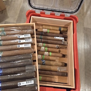 May include: A red, rectangular humidor with a clear lid, filled with cigars. Two wooden trays hold the cigars, some individually wrapped. The cigars vary in color, with green, blue, and white bands. The humidor is on a wooden surface.