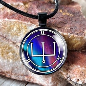 May include: A black necklace with a round pendant featuring a white sigil with a trident symbol on a blue, purple, and pink background. The pendant is surrounded by a black circle with the letters "H", "O", "C", and "O" around the edge.