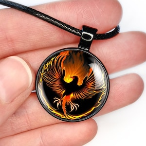 May include: A black necklace with a round pendant featuring a phoenix rising from flames. The phoenix is orange and yellow with black wings and a black body. The pendant is set against a black background.