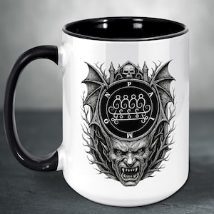 May include: White ceramic mug with a black handle and rim. The mug features a detailed black and white illustration of a demonic face, wings, and sigils. The design is centered on the mug's surface.