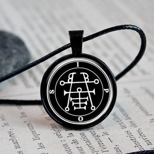May include: A black necklace with a round pendant featuring a white sigil on a black background. The sigil is surrounded by a circle with the letters "I", "B", "O", "P", and "S" around it.