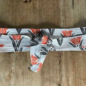 May include: A white fabric headband with a black and orange floral print. The headband is tied in a knot at the front.