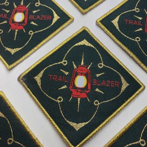 May include: A dark green square patch with gold stitching and a red and white lantern in the center. The words "TRAIL BLAZER" are written in red on the patch.