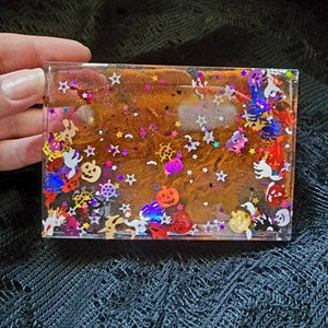 Op de afbeelding: Een transparant harsbakje met een glinsterende oranje-bruine achtergrond. Het bakje is gevuld met verschillende Halloween-charms, waaronder pompoenen, sterren, geesten en spinnen.