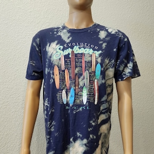 May include: A blue tie-dye t-shirt with a graphic of the evolution of the surfboard. The graphic includes a timeline of surfboard designs from ancient times to the 1970s. The text "Evolution of the Surfboard" is printed in a white and green font. The text "So Cal" is printed in a white font at the bottom of the graphic.