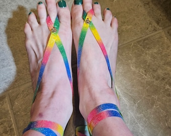 May include: A pair of rainbow-colored sandals with green toe rings. The sandals are made of a thin, stretchy material and have a criss-cross design.