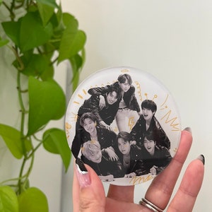May include: A round, clear glass phone grip with a black and white photo of seven people. The photo shows the people in a group pose, with some of them sitting and others standing. The photo is surrounded by a gold outline and includes gold doodles.