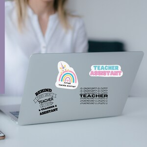 May include: A silver laptop computer with three stickers. The stickers are a rainbow with a unicorn, a black and white text sticker that says "Behind every great teacher is a great teacher assistant", and a pink and blue sticker that says "Teacher Assistant".