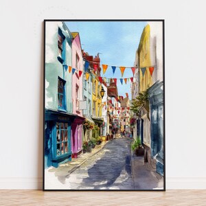 May include: Watercolour painting of a European street scene with colourful buildings and a cobbled road. Buildings are painted in blue, pink, yellow, and green. Strings of triangular flags hang overhead. The artwork is framed in a black frame.