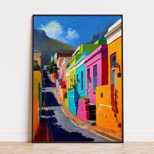 Peut inclure: Une impression d'art encadrée représentant une scène de rue colorée. Des bâtiments aux couleurs orange, rose, vert et bleu bordent une route en montée. Une montagne et un ciel bleu nuageux sont en arrière-plan. L'œuvre est de style impressionniste.