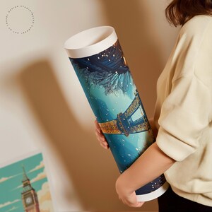 May include: A rolled art print depicting the Eiffel Tower at night, in shades of blue and gold. The print has white end caps and a border. The text "TRAVEL DESIGN STUDIO-BASED ART OF EUROPE" is printed on the side. The print is held by a person.