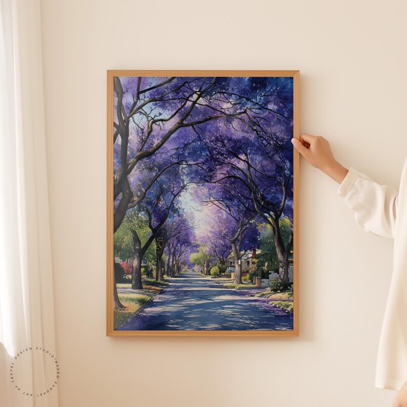 Peut inclure: Une impression d'art encadr&eacute;e repr&eacute;sente une rue bord&eacute;e d'arbres en pleine floraison, cr&eacute;ant une canop&eacute;e de fleurs violettes. L'&oelig;uvre d'art pr&eacute;sente un style aquarelle avec un &eacute;clairage et des ombres doux, am&eacute;liorant la profondeur de la sc&egrave;ne. Le cadre est de couleur bois clair.