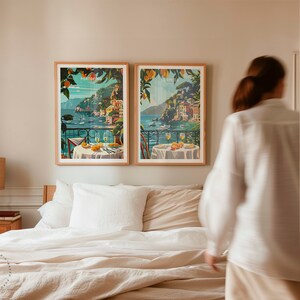 K&ouml;nnte beinhalten: Zwei gerahmte Vintage-Reiseposter mit einer K&uuml;stenlandschaft von Positano und einer Tischdekoration mit Gl&auml;sern und Fr&uuml;chten. Die Poster h&auml;ngen an einer Wand &uuml;ber einem Bett mit wei&szlig;en Bettw&auml;sche und einem braunen Kopfteil.