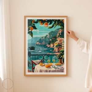 K&ouml;nnte beinhalten: Ein gerahmtes Poster mit einer farbenfrohen Illustration einer K&uuml;stenstadt mit Blick auf das Meer und einem Tisch mit zwei Gl&auml;sern Wein und Orangen. Das Poster tr&auml;gt den Text "PORTOFINO" in wei&szlig;en Buchstaben auf blauem Hintergrund.