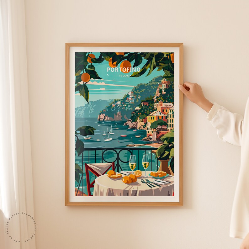 K&ouml;nnte beinhalten: Ein gerahmtes Poster mit einer farbenfrohen Illustration einer K&uuml;stenstadt mit Blick auf das Meer und einem Tisch mit zwei Gl&auml;sern Wein und Orangen. Das Poster tr&auml;gt den Text "PORTOFINO" in wei&szlig;en Buchstaben auf blauem Hintergrund.