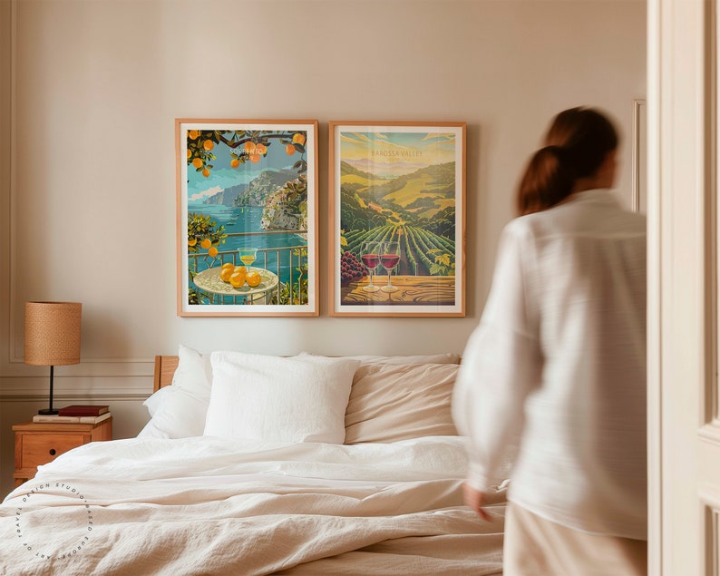 May include: Two framed vintage travel posters, one depicting a coastal scene with a lemon tree and the other showing a vineyard landscape. The posters are hanging on a wall above a bed with white linens.