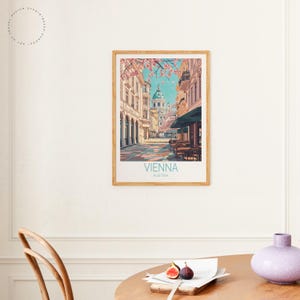 May include: A framed art print depicting a street scene in Vienna, Austria. The print shows a view down a cobbled street towards a domed building. Pink blossoms frame the top of the image, with the words "Vienna" and "Austria" printed at the bottom.