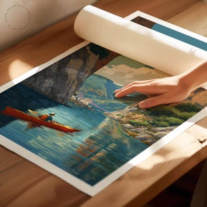 K&ouml;nnte beinhalten: Eine gerollte Kunstdruck zeigt einen Kajakfahrer auf einem See, mit einer Berg- und Klippenlandschaft. Der Druck wird auf einer Holzoberfl&auml;che ausgerollt und gibt das Kunstwerk frei.