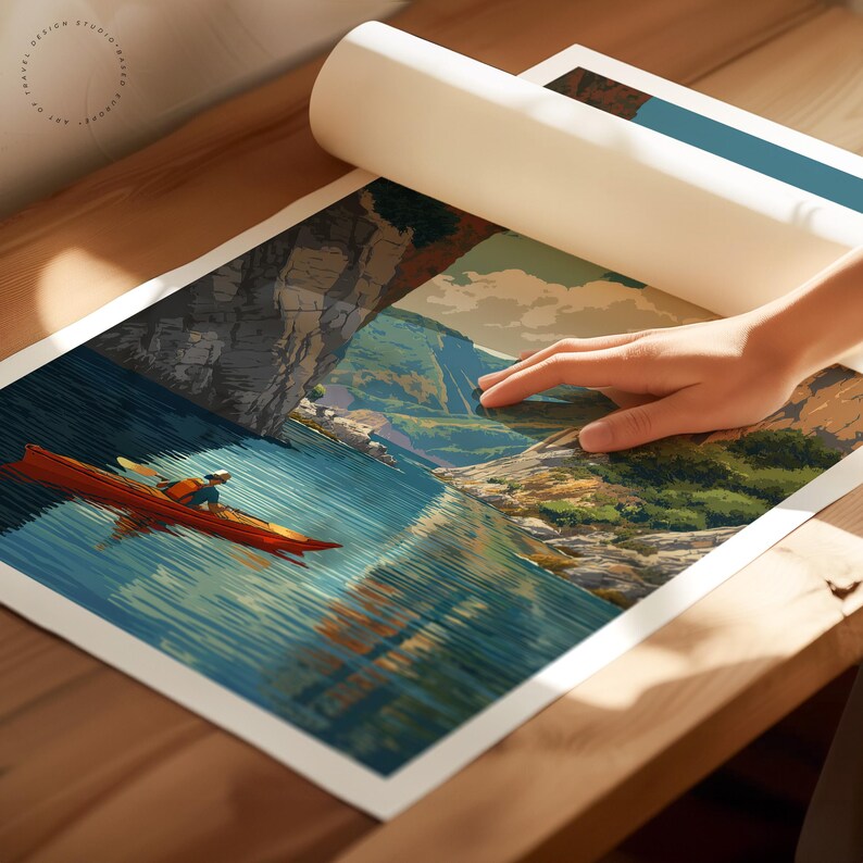 K&ouml;nnte beinhalten: Eine gerollte Kunstdruck zeigt einen Kajakfahrer auf einem See, mit einer Berg- und Klippenlandschaft. Der Druck wird auf einer Holzoberfl&auml;che ausgerollt und gibt das Kunstwerk frei.