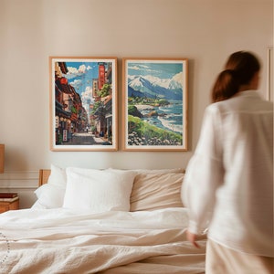 Op de afbeelding: Twee ingelijste prints van Japanse landschappen hangen aan een lichtbeige muur boven een bed met witte lakens. De print aan de linkerkant toont een straatbeeld met gebouwen en mensen die lopen. De print aan de rechterkant toont een bergketen met een strand op de voorgrond.