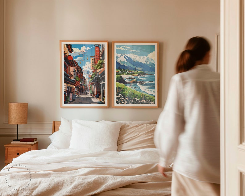 Op de afbeelding: Twee ingelijste prints van Japanse landschappen hangen aan een lichtbeige muur boven een bed met witte lakens. De print aan de linkerkant toont een straatbeeld met gebouwen en mensen die lopen. De print aan de rechterkant toont een bergketen met een strand op de voorgrond.