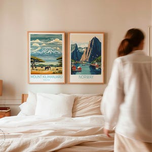 May include: Two framed travel posters on a wall above a bed with white linens. The poster on the left shows a mountain range with the text "Mount Kilimanjaro, Tanzania". The poster on the right shows a fjord with the text "Norway".